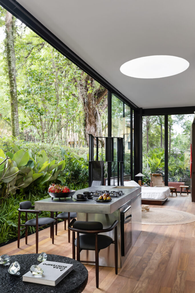 Cozinha da Casa Limiar R com ilha, cooktop, claraboia circular e esquadrias de vidro integrando o estar ao jardim
