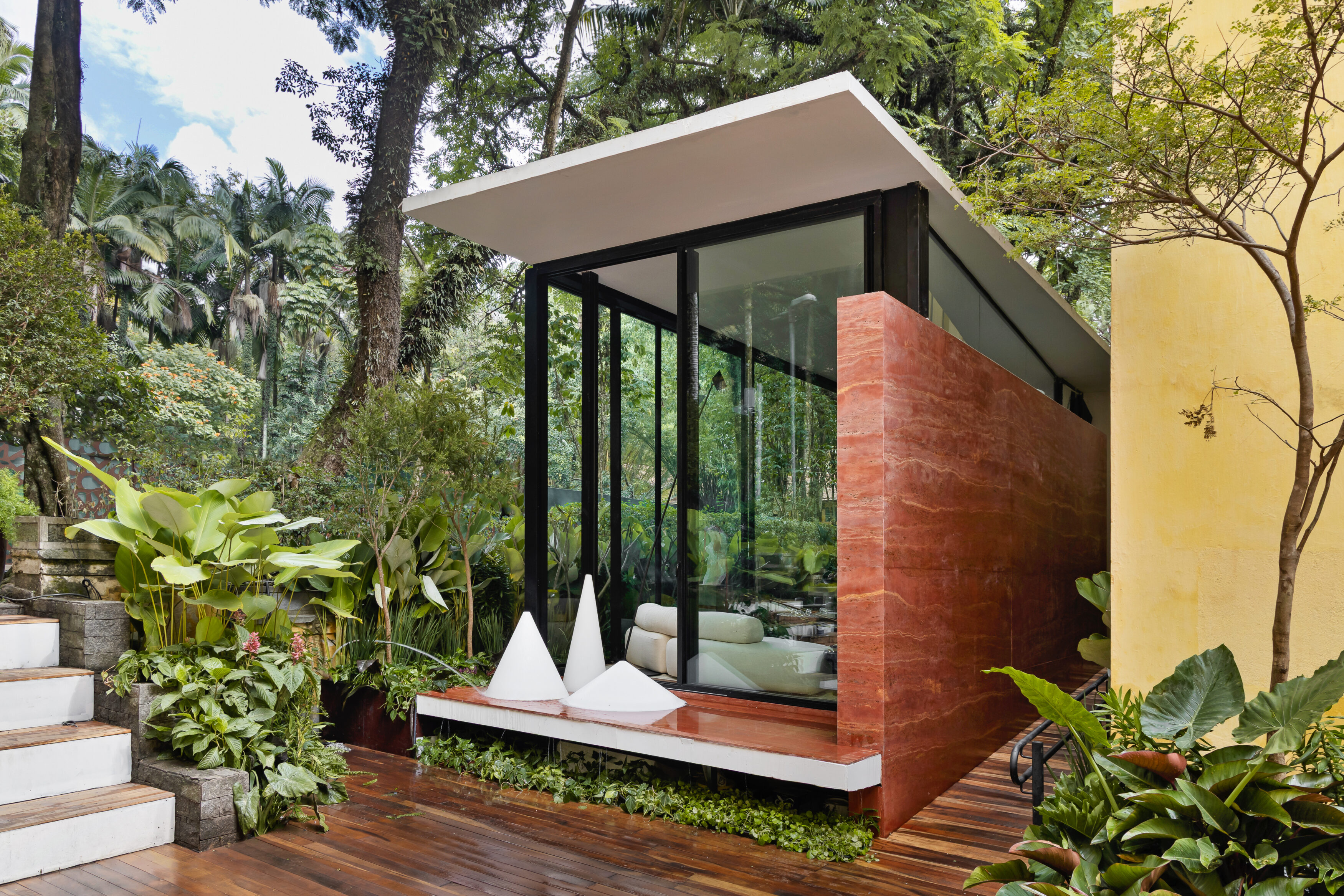 Vista externa da Casa Limiar R com volume envidraçado, deck de madeira e jardim ao redor