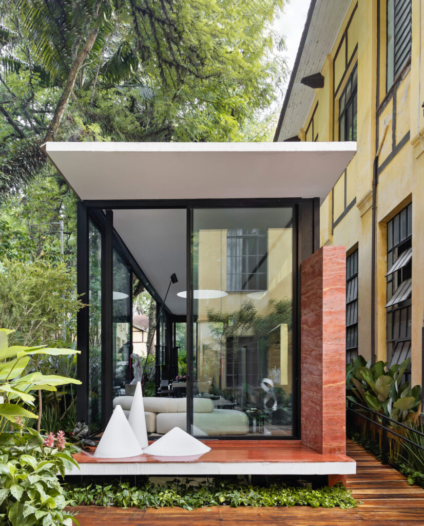 Fachada envidraçada da Casa Limiar R na CASACOR São Paulo 2026, com volume em vidro e parede em Travertino Rosso entre os casarões do Parque da Água Branca