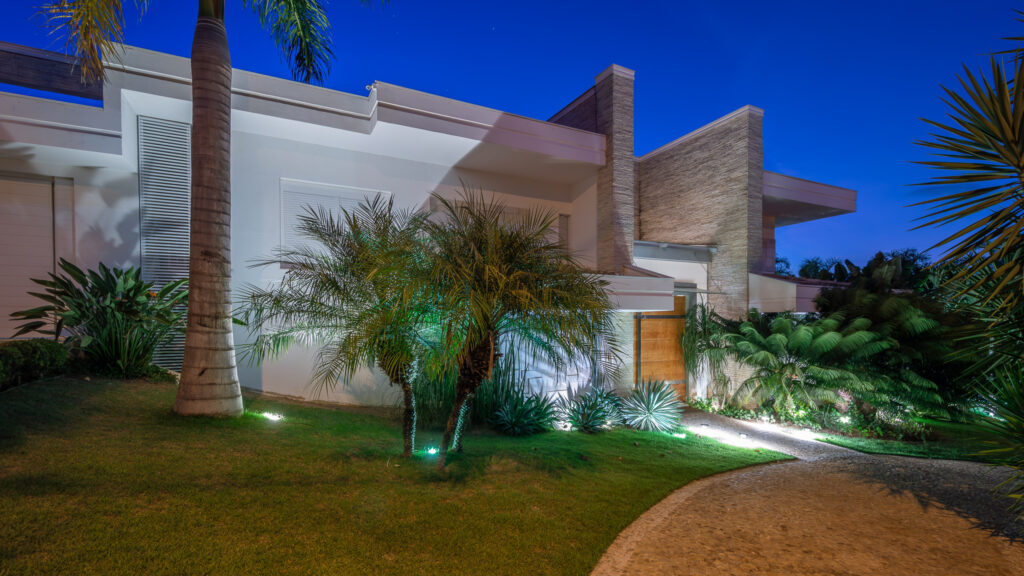 Fachada noturna de residência branca com volume revestido em pedra, porta de madeira e paisagismo tropical iluminado