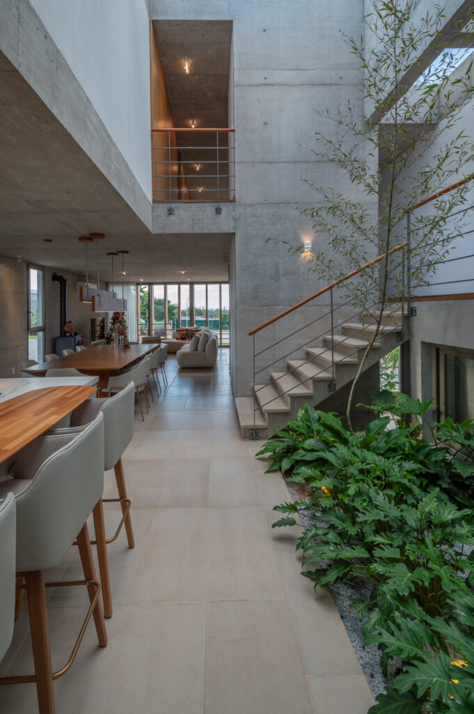 Circulação de pé-direito triplo em concreto aparente junto ao jardim interno, com living e esquadrias Weiku ao fundo