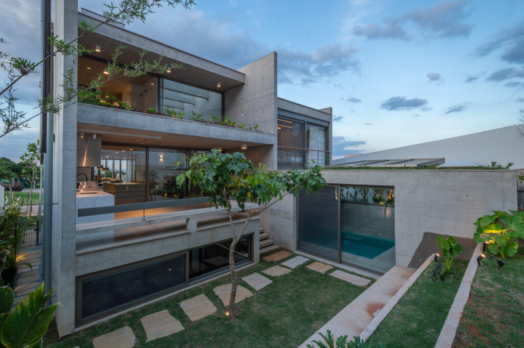 Fachada dos fundos ao entardecer em concreto aparente, com terraços ajardinados, piscina coberta e esquadrias de vidro Weiku iluminadas