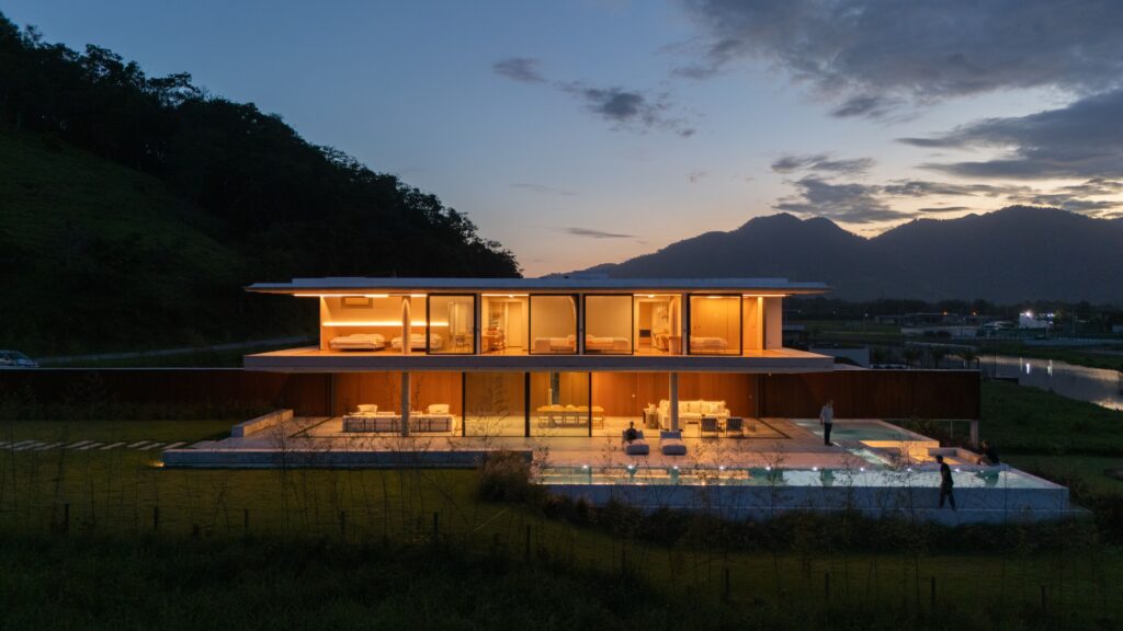Residência em concreto aparente ao anoitecer, com panos de vidro Weiku iluminados nos dois pavimentos e montanhas ao fundo