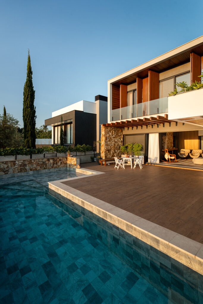 Fachada dos fundos com piscina, deck, parede de pedra e portas de vidro de correr Weiku de perfil preto abertas para o living