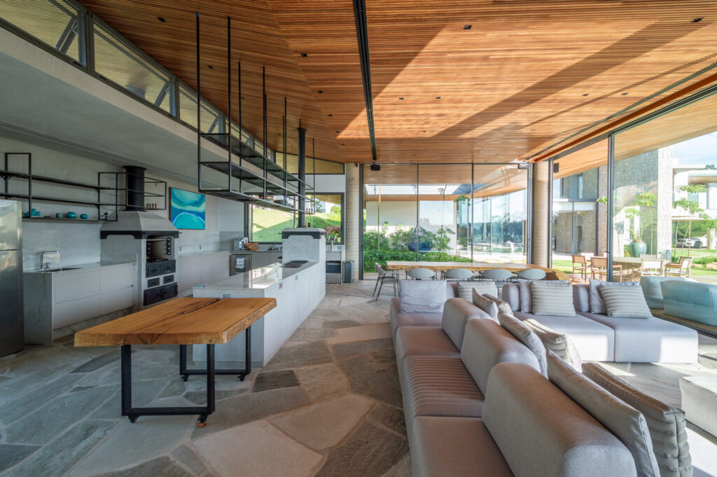 Espaço gourmet com cozinha, churrasqueira e forro de madeira, cercado por panos de vidro Weiku voltados para a piscina