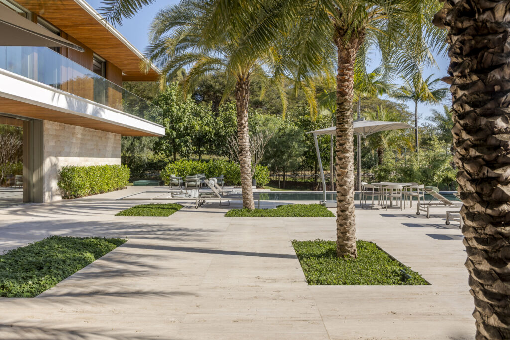 Terraço em travertino com palmeiras e piscina, junto à fachada com forro de madeira e guarda-corpo de vidro