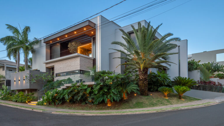 Fachada de esquina de residência contemporânea com revestimento em pedra, volume com forro de madeira iluminado e paisagismo tropical