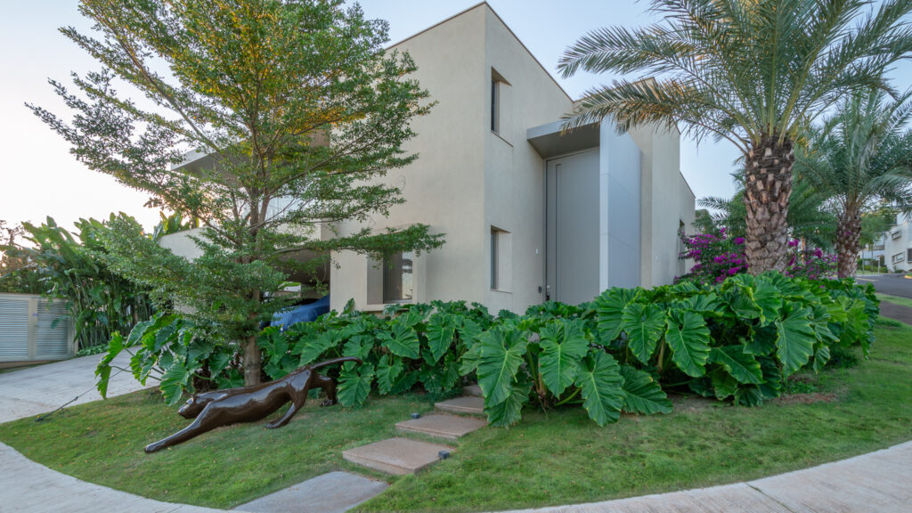 Fachada de esquina em tom areia com porta pivotante, janelas estreitas e paisagismo tropical