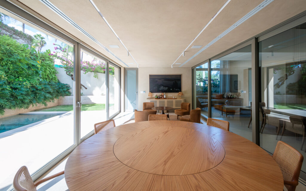 Sala de jantar com mesa redonda envolvida por panos de vidro Weiku de canto voltados para a piscina e o jardim