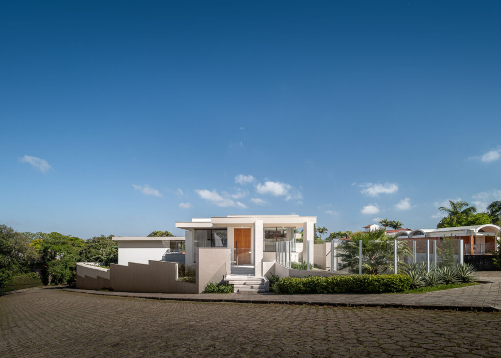 Fachada frontal de residência contemporânea em tons de areia com porta pivotante de madeira e panos de vidro Weiku
