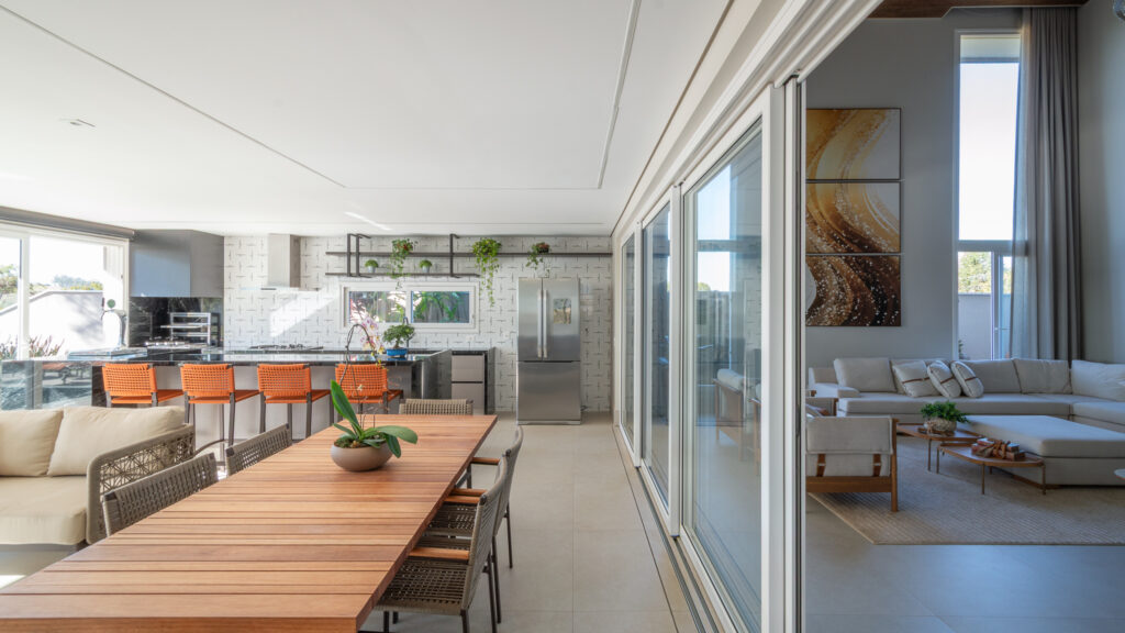 Espaço gourmet com mesa de madeira e cozinha, separado do living por portas de vidro de correr Weiku de perfil branco