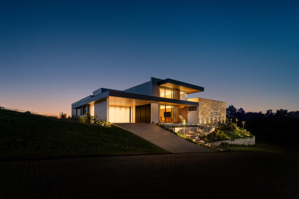 Fachada de residência contemporânea ao entardecer, com parede de pedra iluminada e esquadrias de vidro Weiku