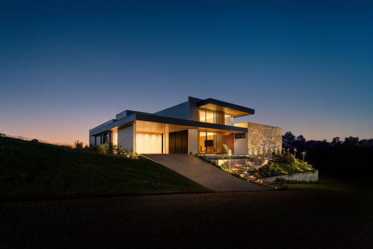 Fachada de residência contemporânea ao entardecer, com parede de pedra iluminada e esquadrias de vidro Weiku