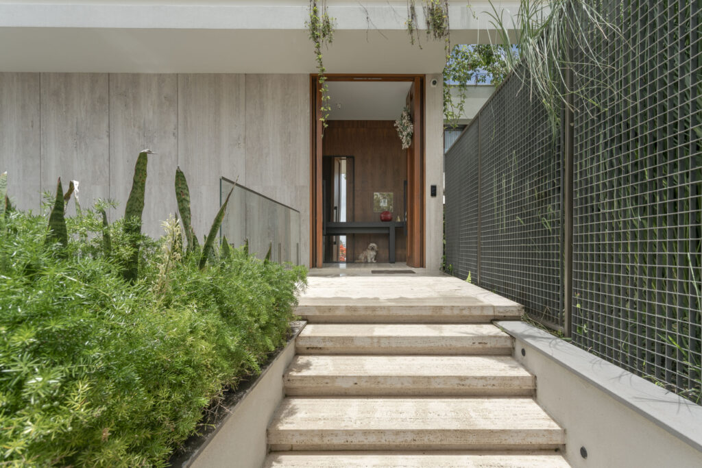 Escada de entrada em travertino com jardim lateral, conduzindo à porta pivotante de madeira da residência