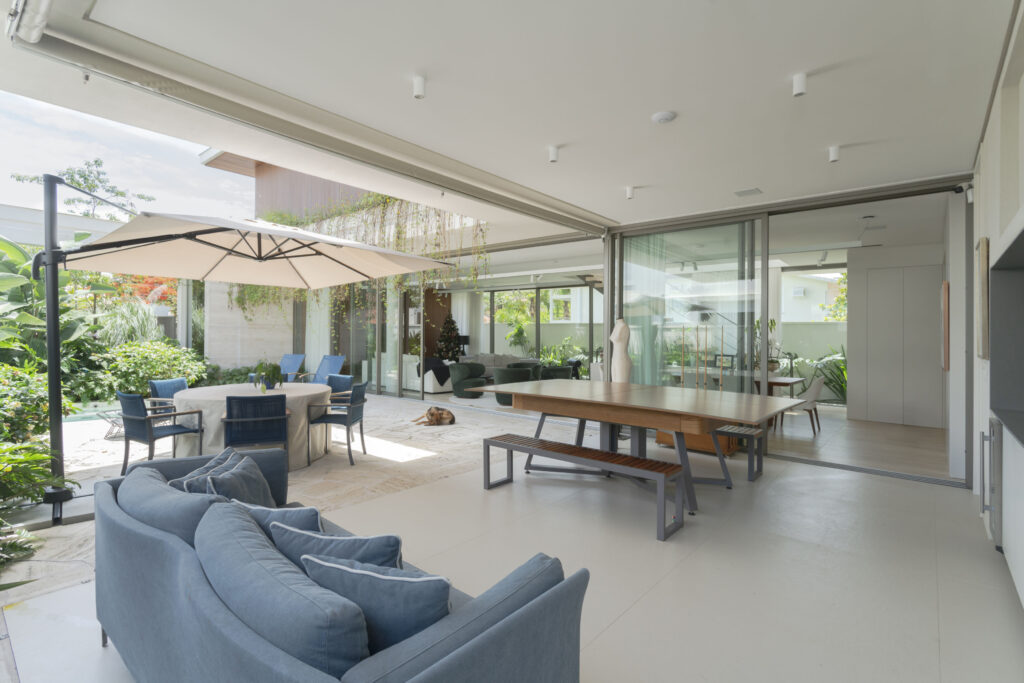 Varanda gourmet integrada ao living por portas de vidro de correr Weiku de perfil escuro, abertas para o jardim