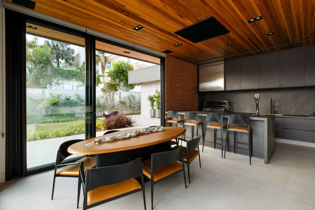 Espaço gourmet com cozinha em pedra escura, mesa oval e portas de vidro de correr Weiku de perfil preto voltadas para o jardim